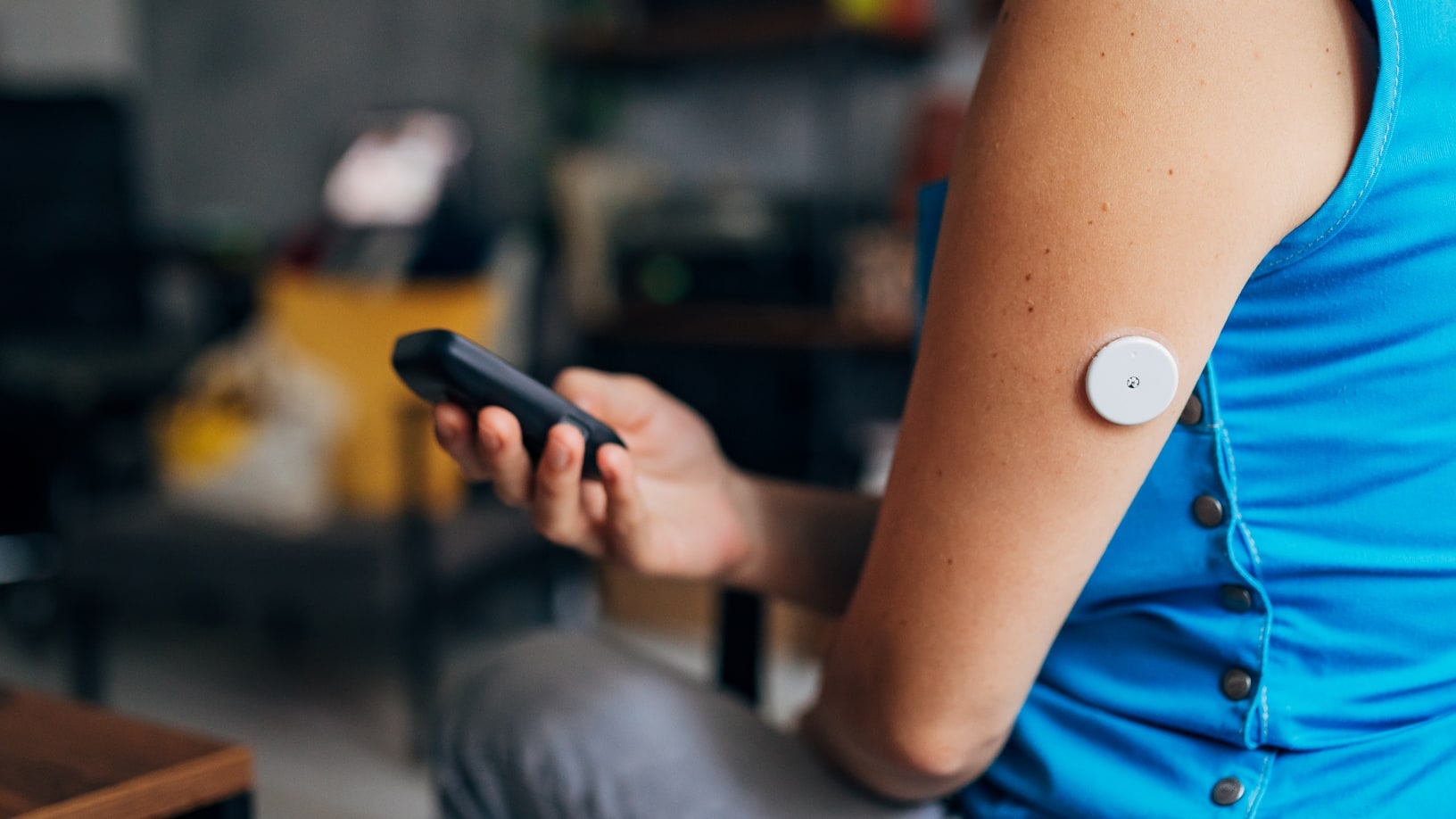 A close-up of a person's upper arm with a continuous glucose monitor sensor, while they hold a remote control or small device in their hand