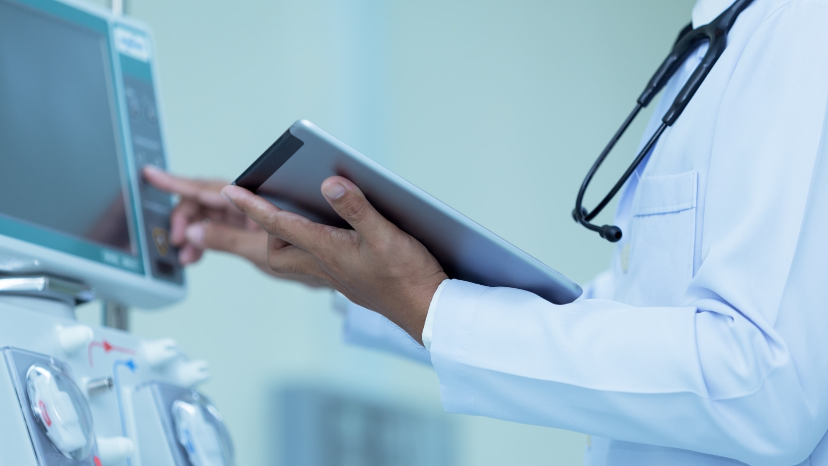 A medical professional in a lab coat and stethoscope interacting with a large medical device, likely a dialysis machine, while holding a tablet