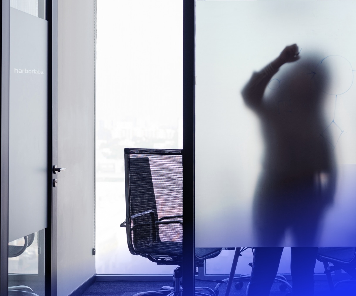 Silhouetted figures of business people behind a frosted glass wall in a modern office, one appearing to write on the glass.