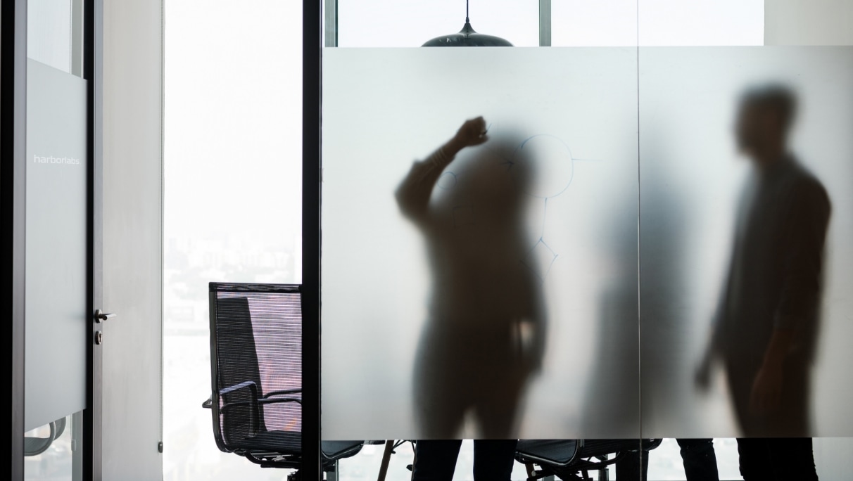 Silhouetted figures of business people behind a frosted glass wall in a modern office, one appearing to write on the glass
