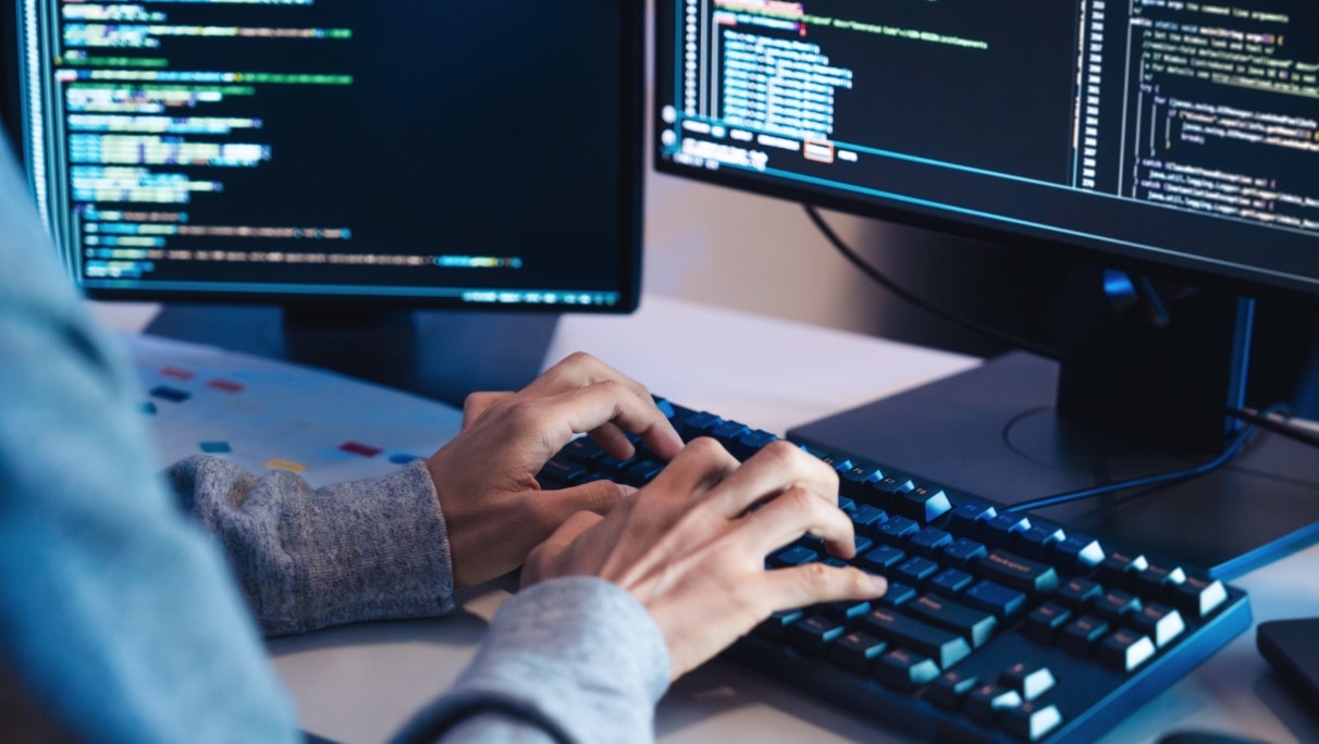 programming coding cybersecurity Close-up of hands typing on a keyboard in front of dual monitors displaying lines of code.