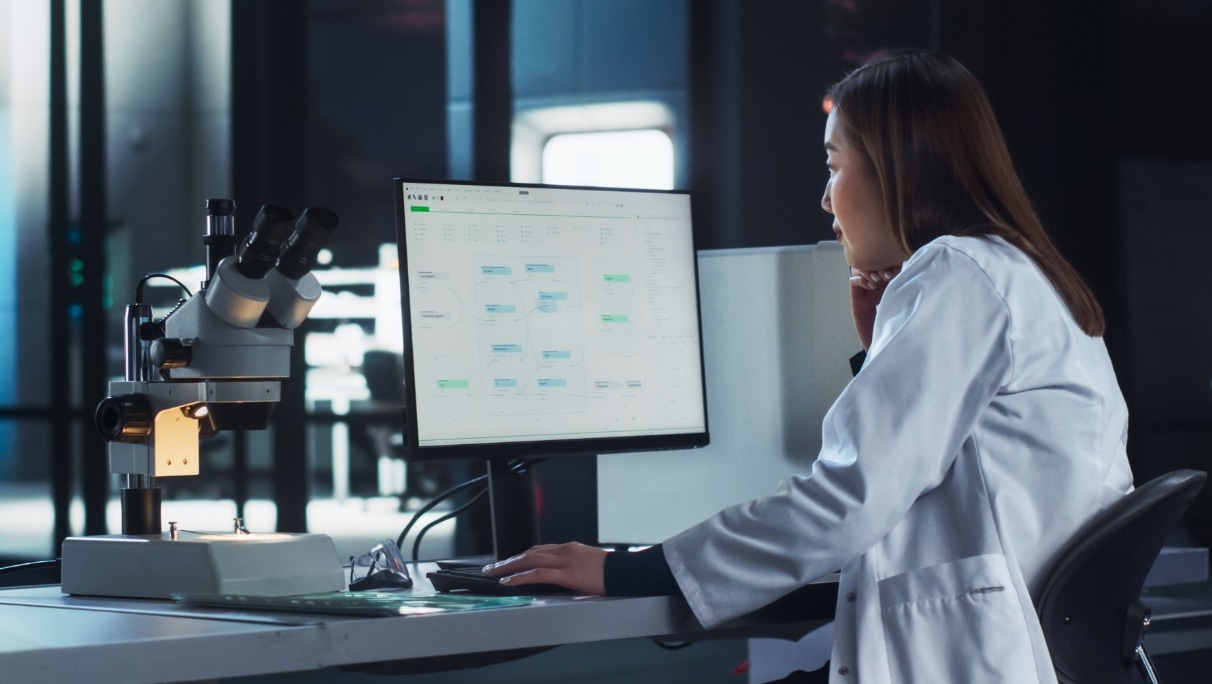 scientist computer schedule A female scientist in a lab, looking at a computer monitor displaying project schedule, with a microscope on the left.
